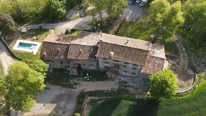 An aerial view of a stone building with a tiled roof, surrounded by greenery and a swimming pool.