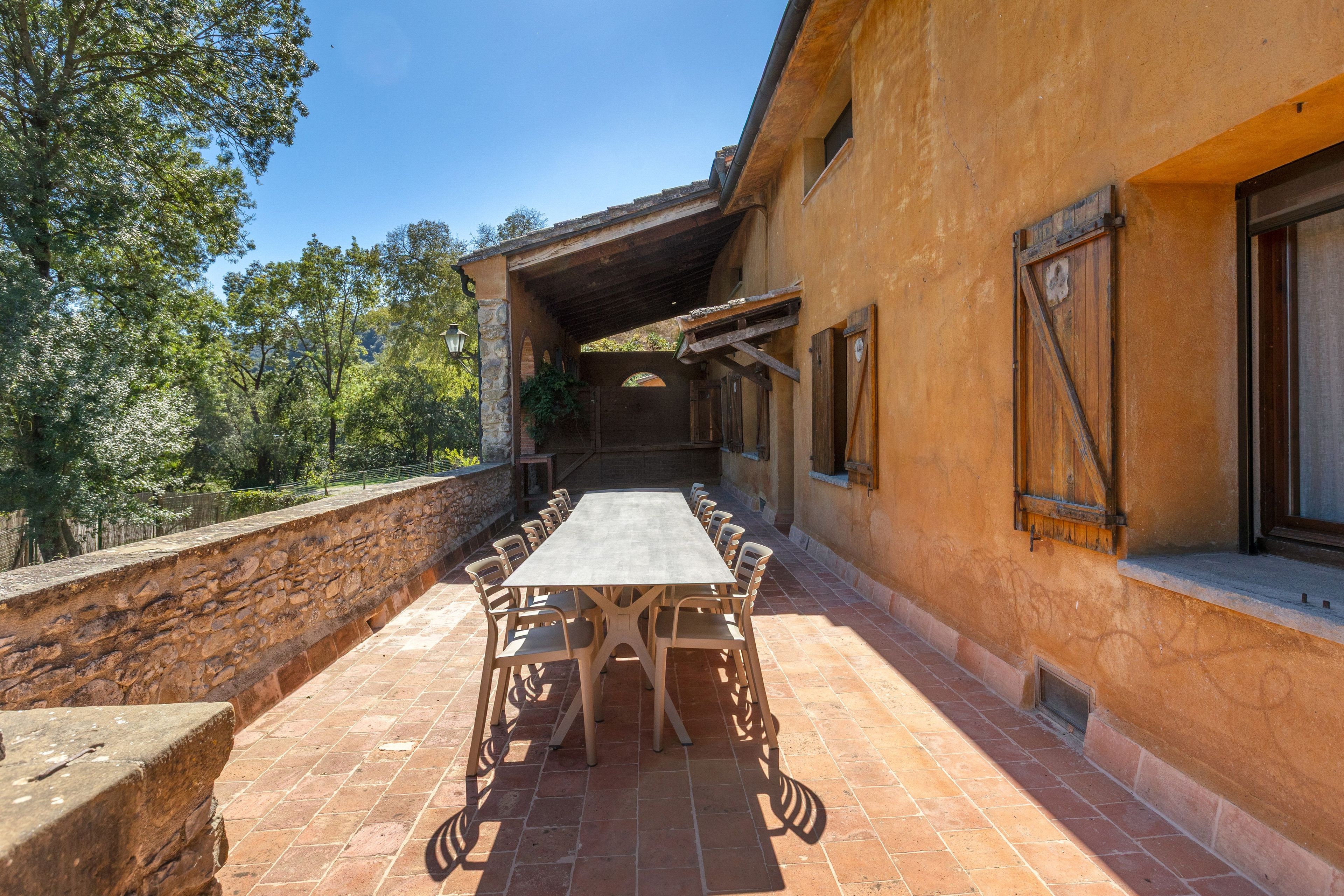 Un patio al aire libre con una mesa larga y sillas, un muro de piedra y un edificio con contraventanas de madera.