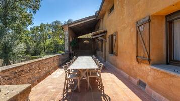 Un patio al aire libre con una mesa larga y sillas, un muro de piedra y un edificio con contraventanas de madera.