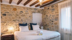 A bedroom with a stone wall, a bed with white bedding, a nightstand, a bedside lamp, and a vase with flowers.