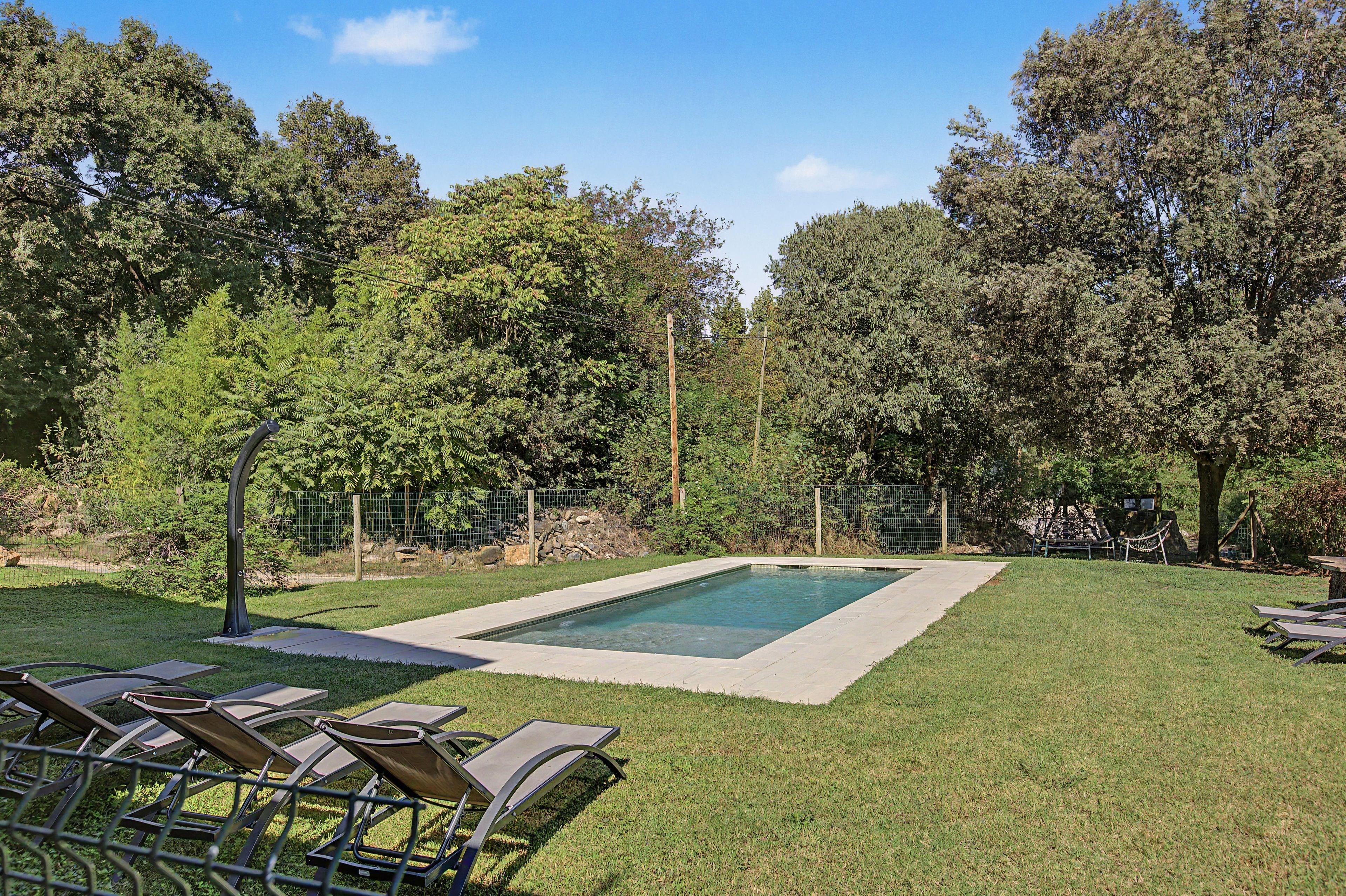 Piscina rectangular rodeada por un césped con árboles, una valla y sillas de descanso.