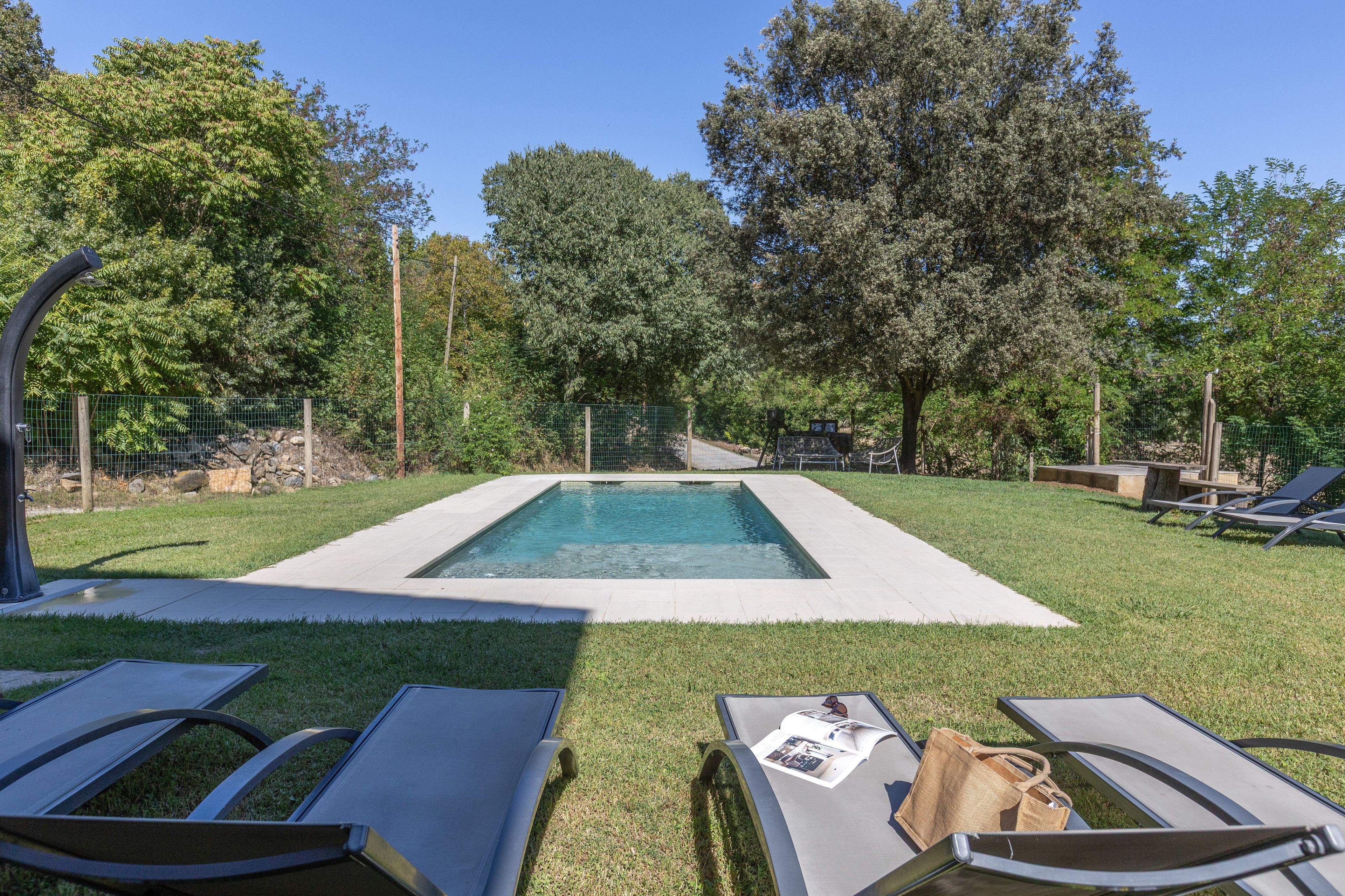 Una piscina rectangular con un césped circundante, dos sillas de descanso y una valla de madera.