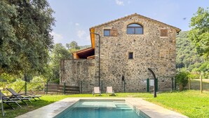 A stone building with a swimming pool, surrounded by greenery and a fence.