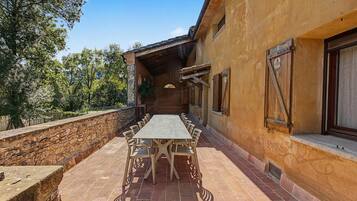 A covered outdoor dining area with a long table and chairs, stone walls, and a view of trees and mountains.