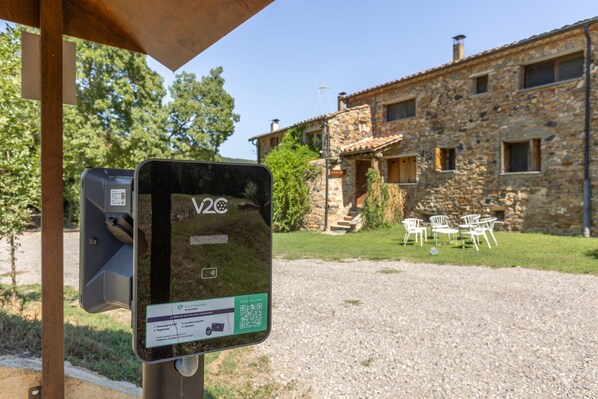 Una estación de carga V2C con instrucciones, un edificio de piedra con tejado de baldosas al fondo y zona de estar exterior.