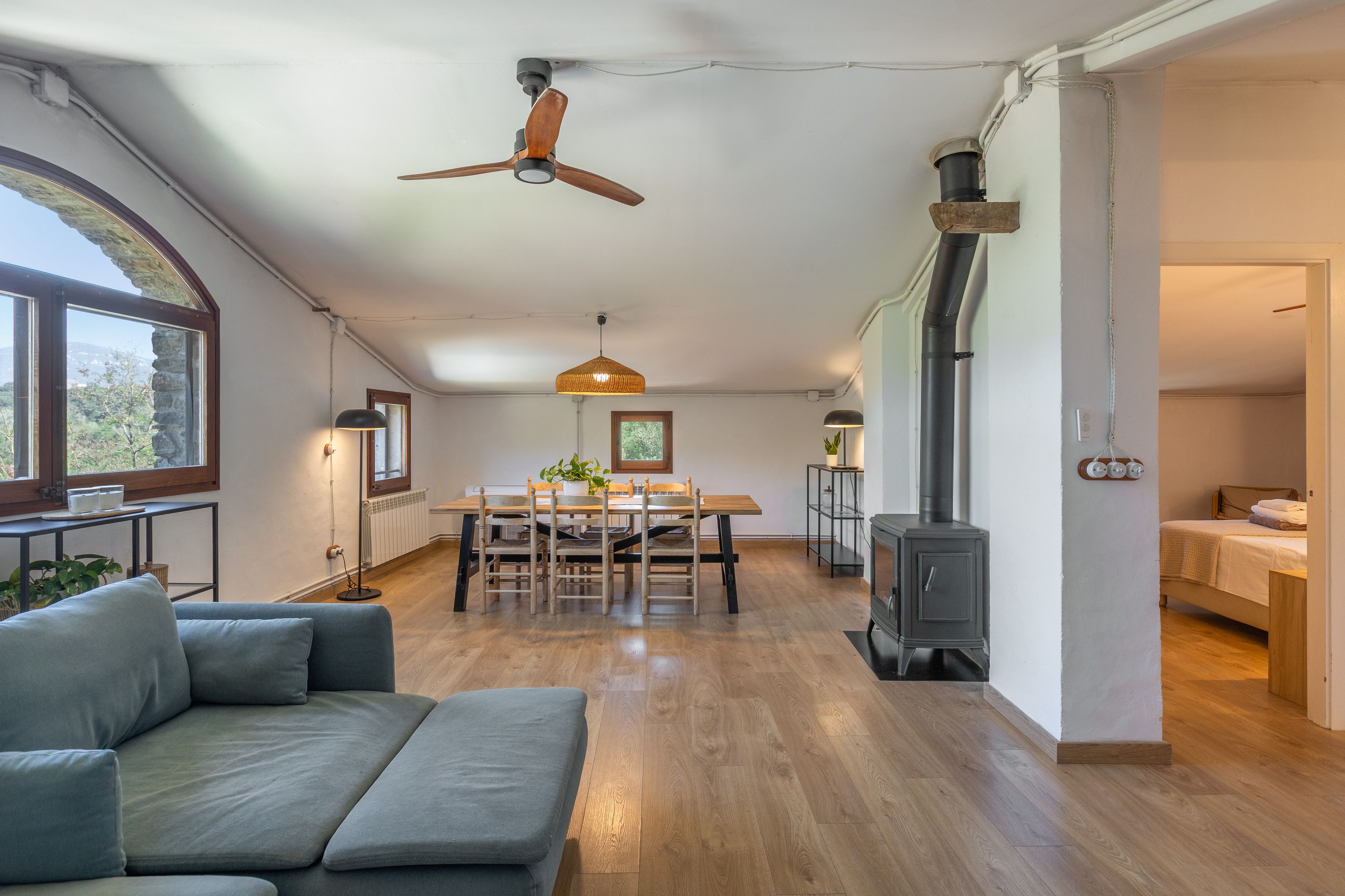Amplia sala de estar con piso de madera, una mesa de comedor con sillas, un sofá, ventilador de techo, calentador de pared y estufa de leña.