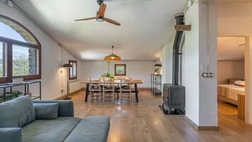 Amplia sala de estar con piso de madera, una mesa de comedor con sillas, un sofá, ventilador de techo, calentador de pared y estufa de leña.