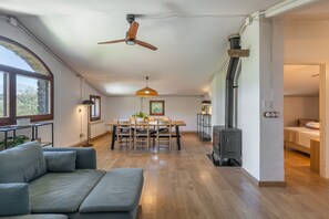 Ein geräumiger Wohnbereich mit Holzboden, einem Esstisch mit Stühlen, einer Couch, einem Deckenventilator, einer Wandheizung und einem Kaminofen.