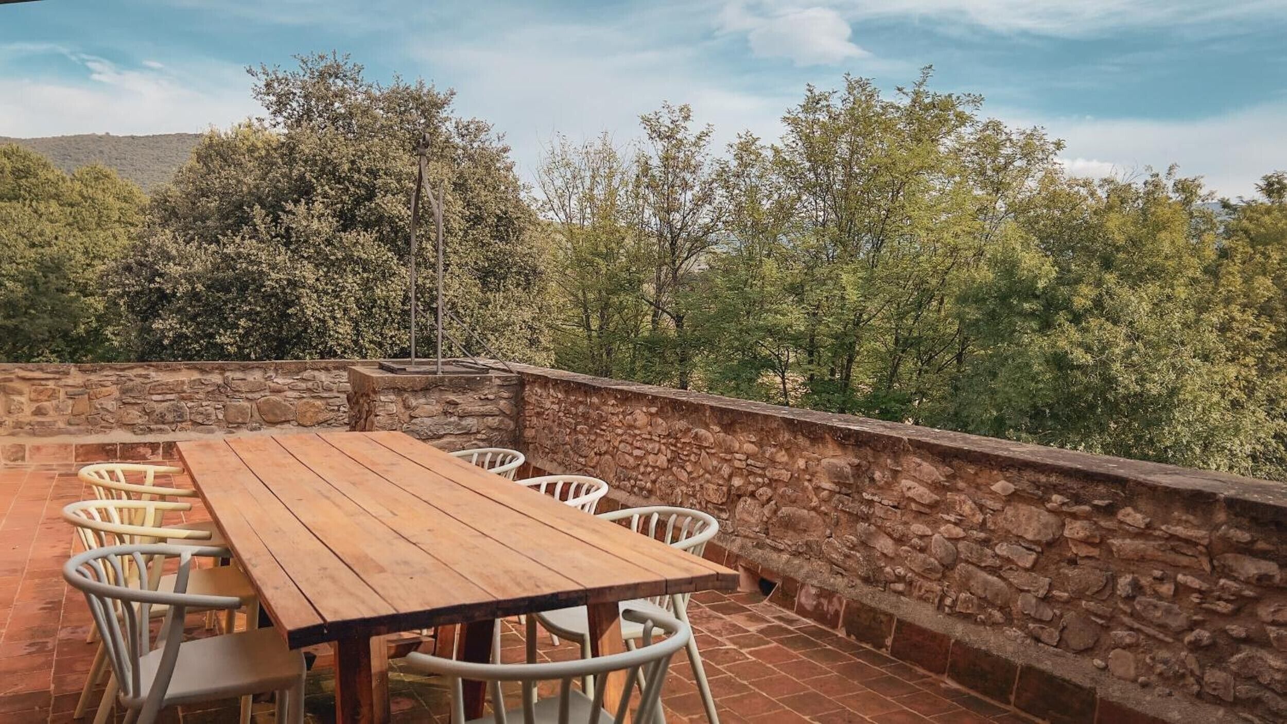 Eine Terrasse im Freien mit Holztisch und weißen Stühlen, umgeben von Steinmauern und Bäumen.