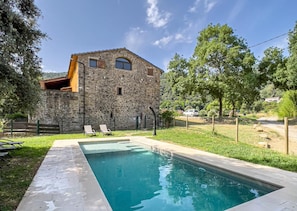 A stone building with a swimming pool, two lounge chairs, and a fenced area.