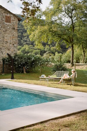 Un edificio de piedra, una piscina y dos sillas de descanso, con un área verde y árboles al fondo.