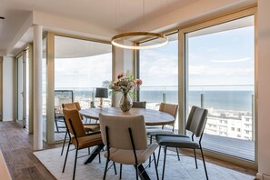 A modern dining area with a round table, chairs, a vase with flowers, and a view of the city and sea through large windows. - High Waves Ostend (Ostend)