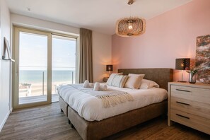 A bedroom with a large bed, a nightstand, a lamp, a painting, and a view of the beach. - High Waves Ostend (Ostend)