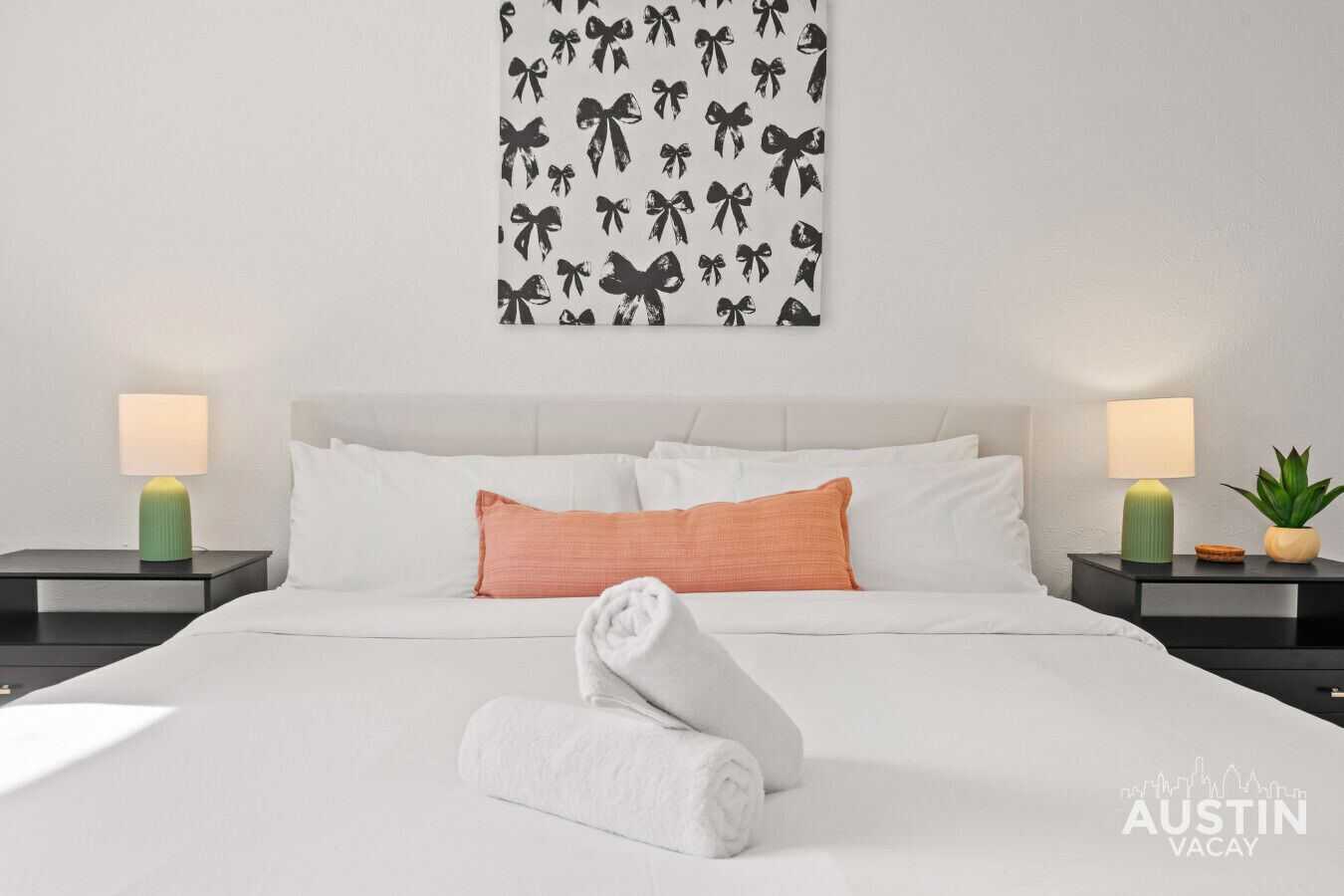 Een hotelkamer met een net opgemaakt bed, twee nachtkastjes met lampen, wanddecoratie met strikken en een watermark van een stadsachtergrond.
