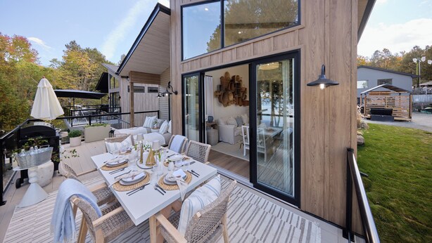 Ett modernt hus med en stor terrass, en utomhusplats för måltider och en glasdörr som leder in till ett vardagsrum.