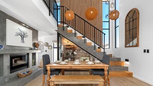 Un comedor con una mesa de madera dispuesta para cuatro, una chimenea y una escalera con barandilla de madera.