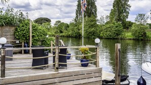 A wooden dock with seating, a flag, and a boat. - River Haven - River Thames Frontage House (Thames Ditton)
