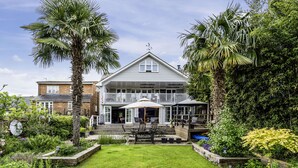 A two-story house with a covered patio, surrounded by lush greenery and a well-maintained lawn. - River Haven - River Thames Frontage House (Thames Ditton)