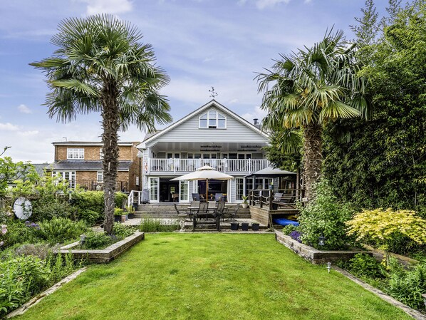 A two-story house with a covered patio, surrounded by lush greenery and a well-maintained lawn. - River Haven - River Thames Frontage House (Thames Ditton)