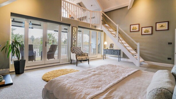 A bedroom with a large bed, a staircase, a balcony with chairs, and a view of the outdoors. - River Haven - River Thames Frontage House (Thames Ditton)