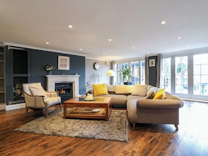 A living room with a fireplace, a sofa set, a coffee table, and a large window providing natural light. - River Haven - River Thames Frontage House (Thames Ditton)