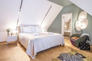 A modern bedroom with a sloped ceiling, a large bed with white linens, bedside lamps, and a decorative wall light. - Bahnhofstrasse Designer Penthouse Private Rooftop (Zürich)