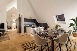 A modern dining area with a dark wooden table set for four, surrounded by light-colored chairs. A kitchen with dark cabinetry is visible in the background, and a flat-screen TV is mounted on the wall. - Bahnhofstrasse Designer Penthouse Private Rooftop (Zürich)