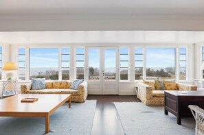 A spacious living room with large windows, a sofa set, a wooden coffee table, and a desk. - Timeless Seaside Estate: 3 Lodgings, Private Beach (Gloucester)