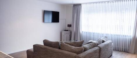 Ein modernes Wohnzimmer mit einer großen Couch, einem flachbildfernseher an der Wand und einem großen Fenster mit durchscheinenden Vorhängen.