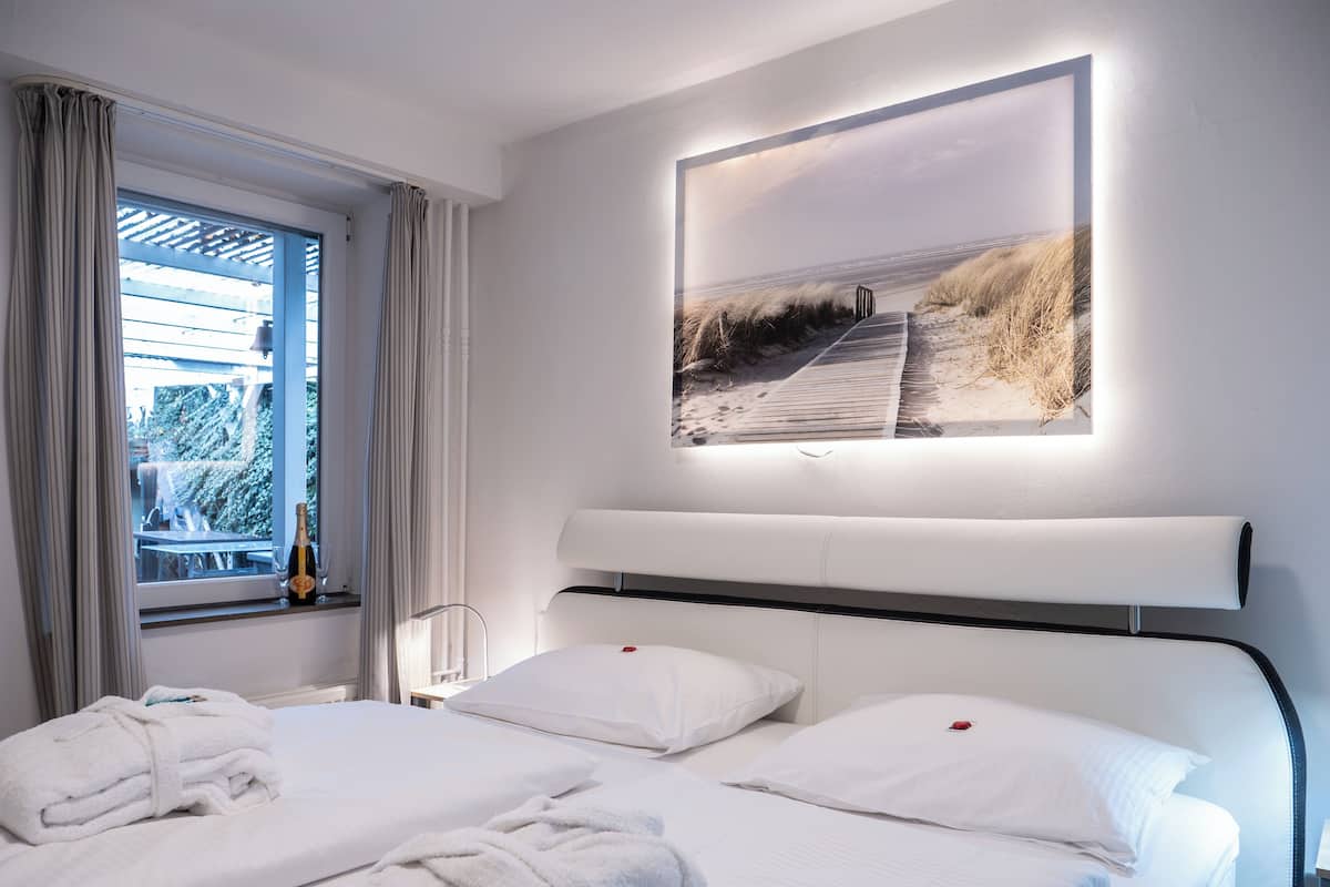 Een hotelkamer met een groot bed, twee kussens, een raam met gordijnen, een nachtkastje met een fles en een ingelijst schilderij aan de muur.