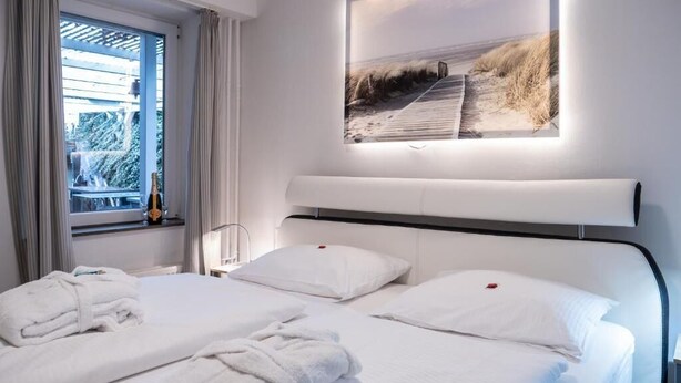 A hotel room with two beds, a large window with curtains, a beach-themed wall art, and a bottle on the windowsill. - Hekmat Apartments Husum Nordsee (Husum)