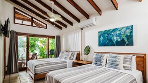 Ett hotellrum med två sängar, en takfläkt, ett stort fönster med gardiner, en tavla på väggen och en krukväxt.