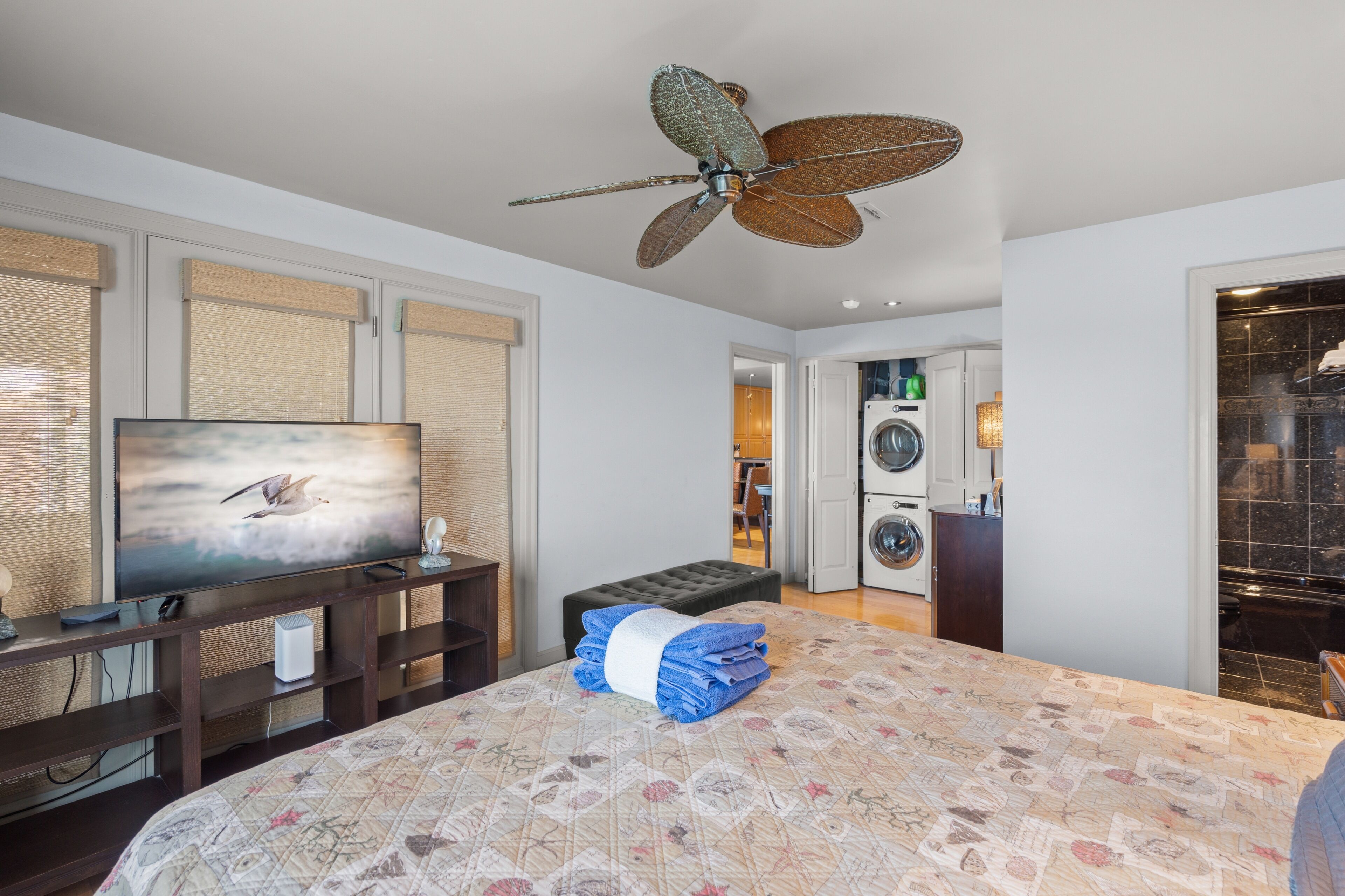 Quarto com cama, televisão, ventoinha no teto, máquina de lavar e banheiro visível ao fundo.