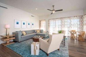 A modern living room with a grey sectional sofa, a round coffee table, a white armchair, and a dining area with a wooden table and wicker chairs.