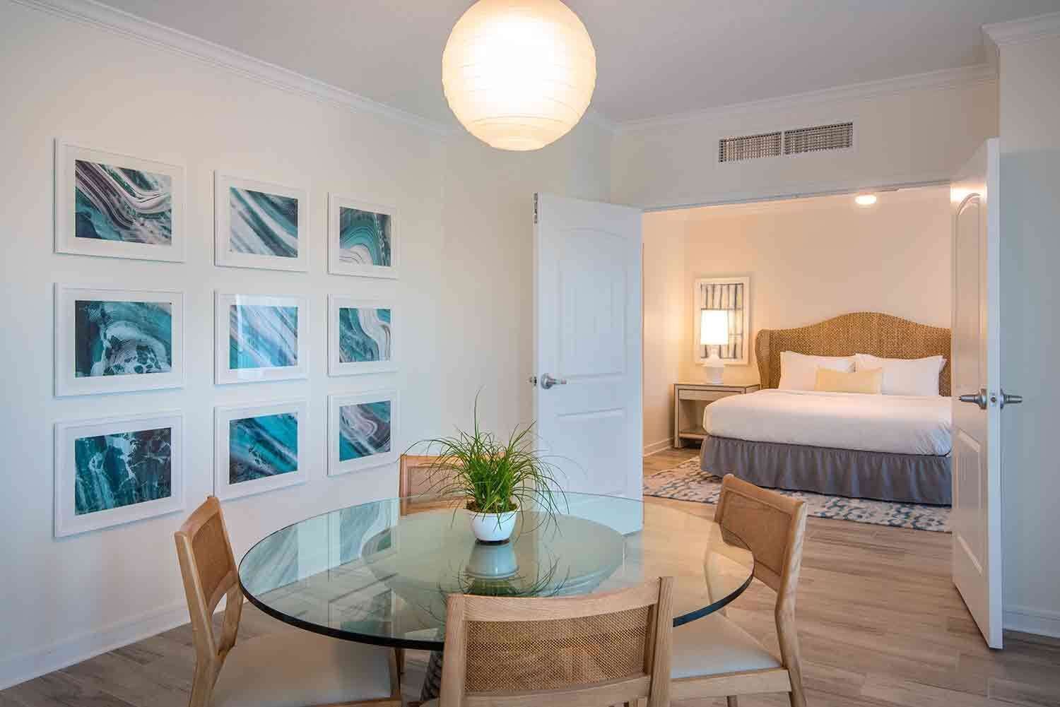 Una camera d'albergo con un tavolo in vetro, sedie, una pianta in vaso, un letto con poggiatesta, un comodino e una parete decorata con quadri.