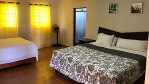 Une chambre d’hôtel avec deux lits simples, un grand lit recouvert d’une couvre-lit à motifs, un ventilateur sur pied et deux tableaux accrochés au mur.