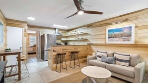 Una cocina moderna con paneles de madera, una isla central con taburetes, una zona de comedor con mesa y sillas, y una sala de estar con sofá y mesa de centro.
