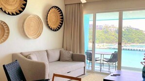Una sala de estar moderna con un sofá, una mesa de comedor y un televisor. Se observan decoraciones de pared tejidas y vistas al mar a través de amplias ventanas.