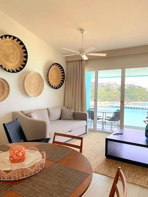 Un salon moderne avec un canapé, une table à manger et un téléviseur. Les murs sont décorés de motifs tissés et de grandes fenêtres offrent une vue sur la mer.