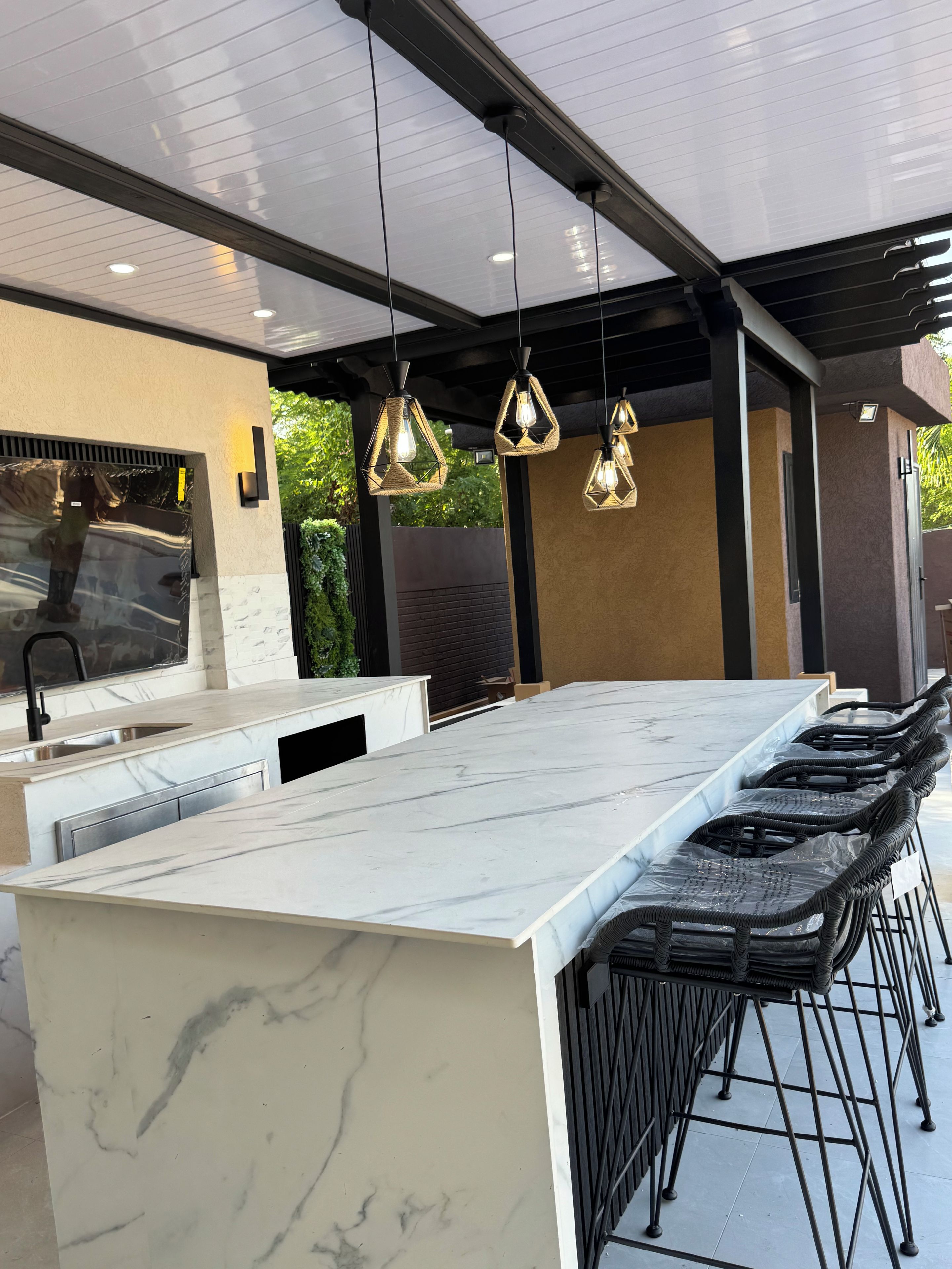 Une cuisine extérieure moderne avec un plan de travail en marbre, des tabourets de bar noirs et des luminaires suspendus.
