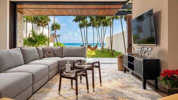 A living room with a sofa, a dining table, chairs, a TV, and a view of the ocean with palm trees.