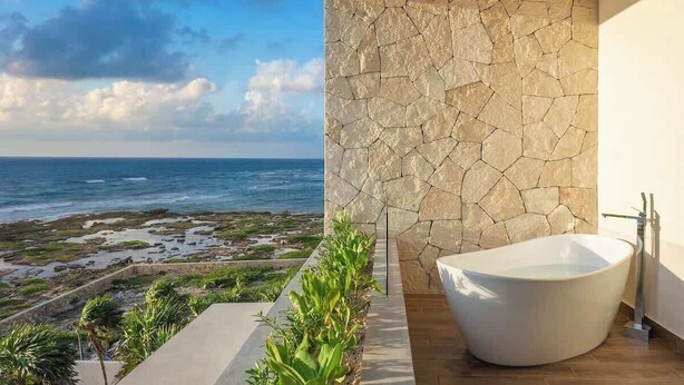 A modern bathroom with a freestanding bathtub, a stone wall, and a view of the ocean. - Valina (Tulsayab)