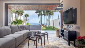 A living room with a sofa, coffee table, and TV, overlooking a beach with palm trees. - Valina (Tulsayab)
