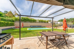 Un espacio al aire libre techado con piscina, una parrilla y una mesa de comedor con sillas.