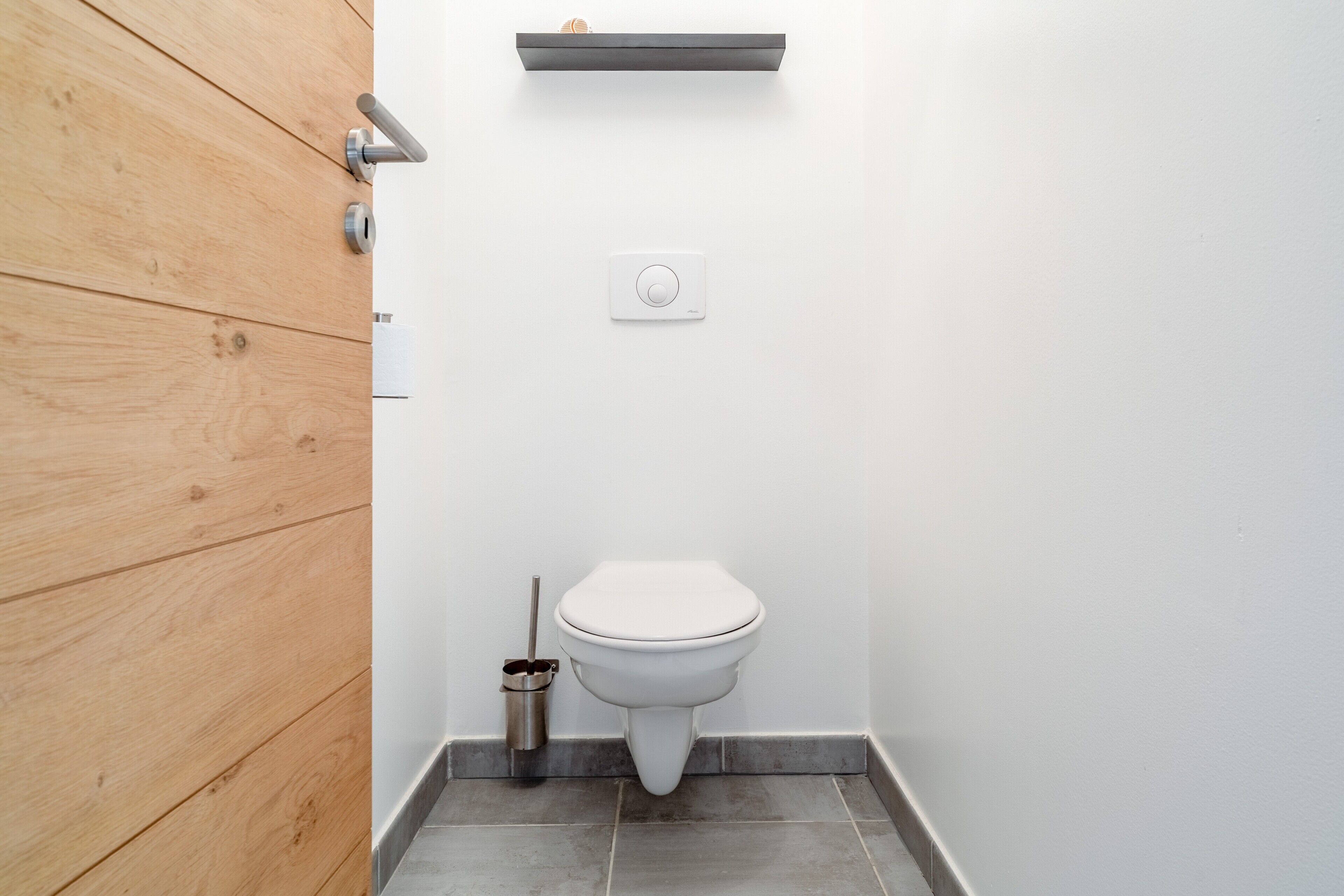 Une salle de bain moderne avec un mur recouvert de panneaux en bois, des toilettes blanches, une poignée de chasse d'eau, un bouton de chasse d'eau et un bidet en métal.