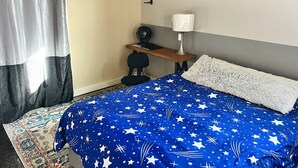 Un dormitorio con cama, escritorio, silla, lámpara y obras de arte en la pared.