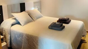 Una cama bien tendida con almohadas y toallas dobladas, un cabecero, una lámpara de pared y un cuadro enmarcado.