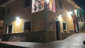 Un edificio de dos pisos con balcón, iluminado por luces, y un área de acceso pavimentada.