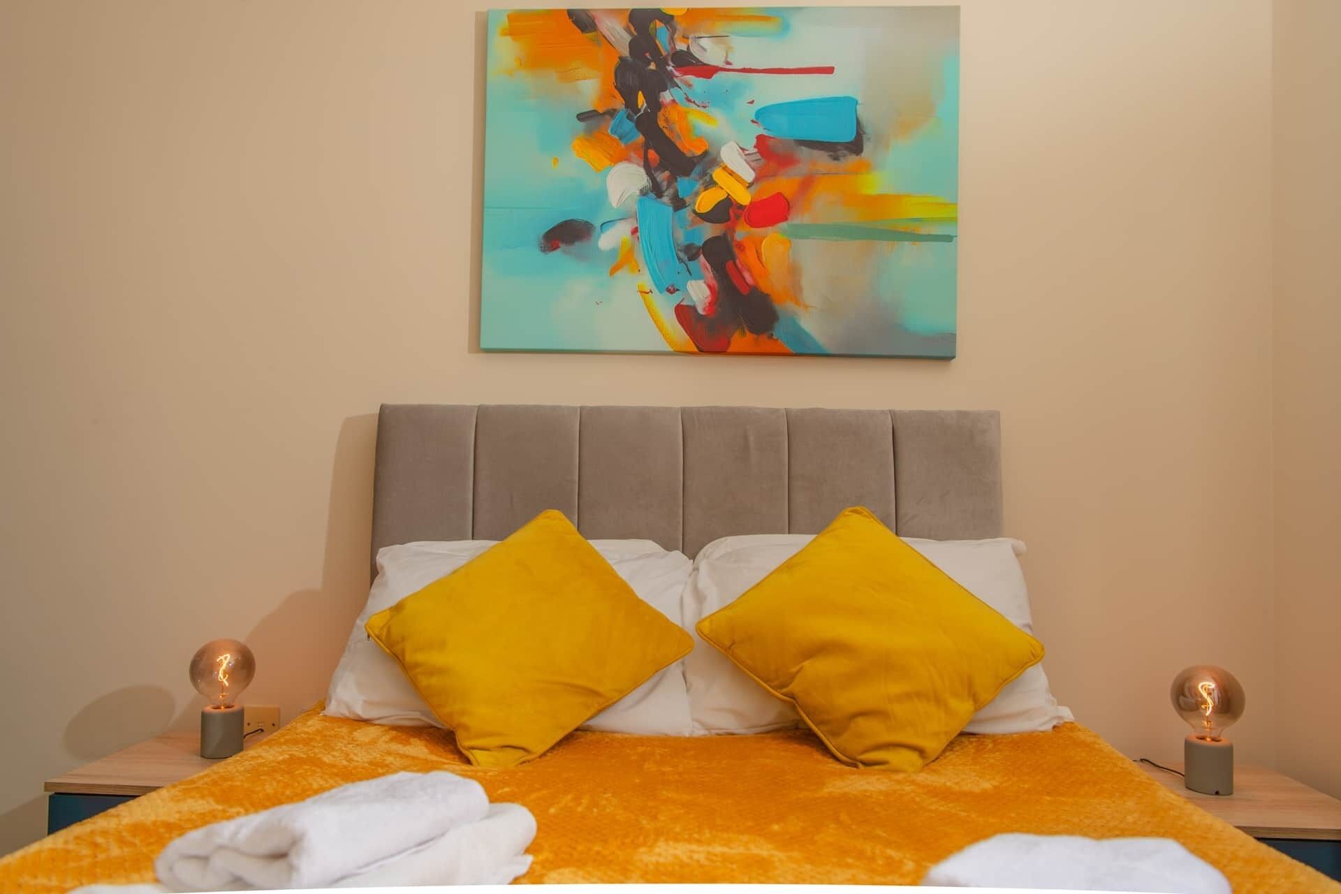 Una cama con dos almohadas amarillas, un cabecero gris y un cuadro abstracto colorido encima.