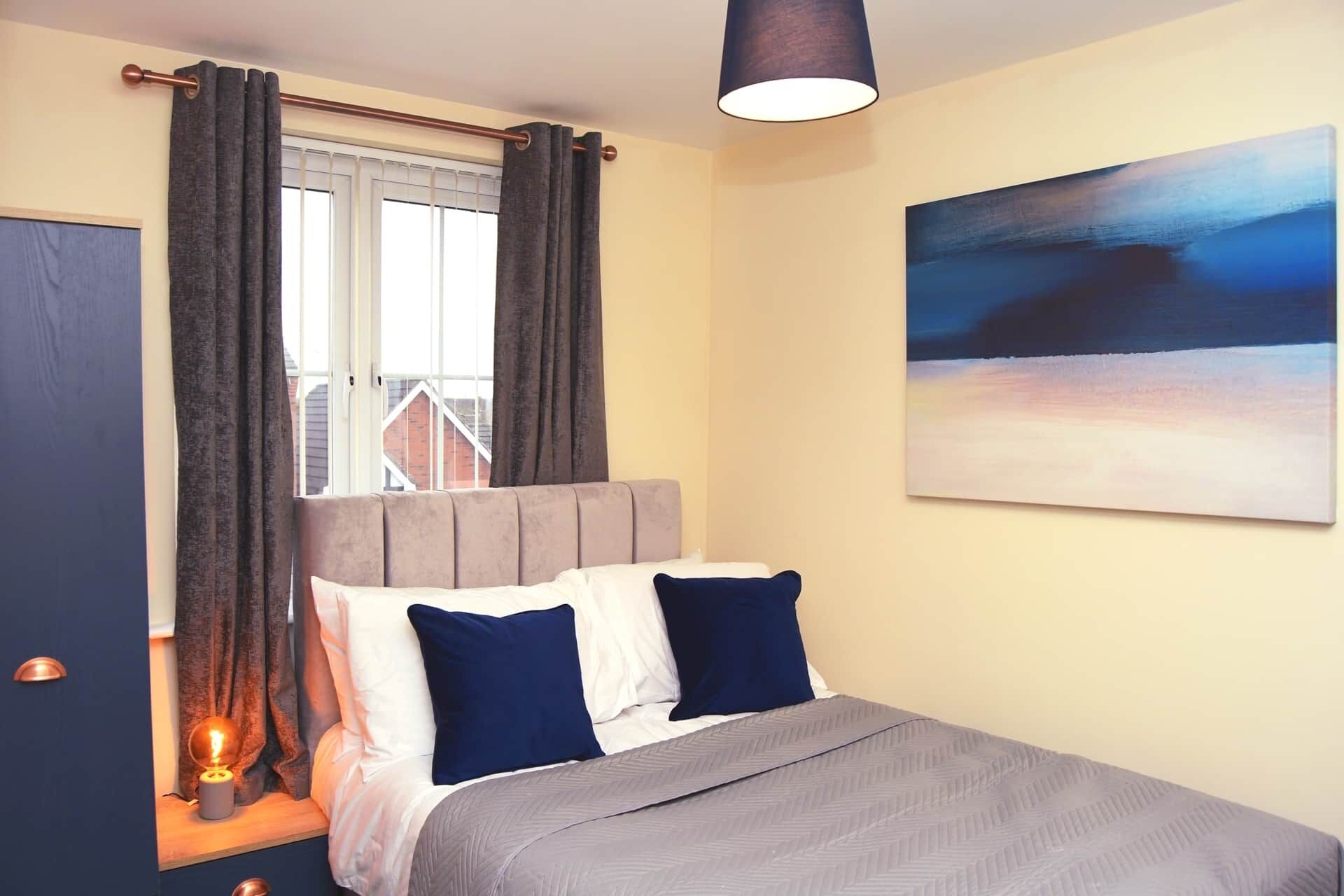 A bedroom with a bed, curtains, a painting, and a lamp.